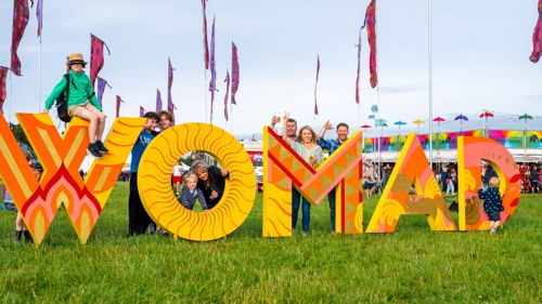 People are standing behind, on and sitting in a series of large colourful letters that spell out WOMAD on a festival site on a sunny day