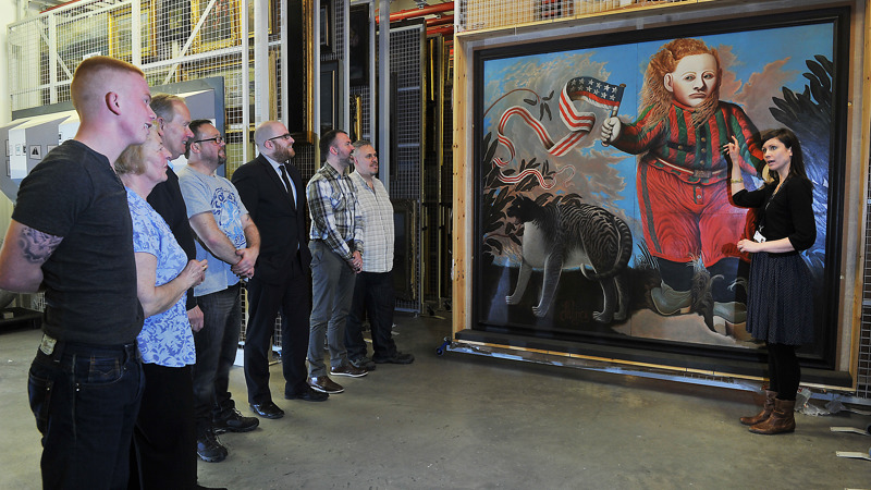A curator talks about a painting to a group of visitors. The painting shows a red haired boy, wearing red clothes waving a distorted American flag.