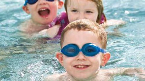 Three children in a swimming pool, two are wearing swimming goggles and all are smiling at the camera