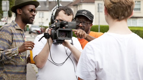 Two adults give advice to a young person with a film camera, who is filming another young person. 
