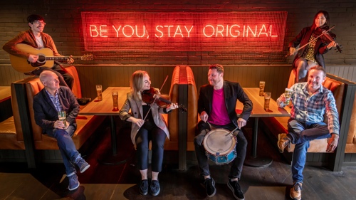 Four members of Scottish trad band Astro Bloc with their instruments, two have fiddles, one has a drum and the other a guitar. They are sitting inside a pub and are joined by Donald Shaw Creative Producer of Celtic Connections and Dougal Sharp Master Brewer and founder of Innis & Gunn