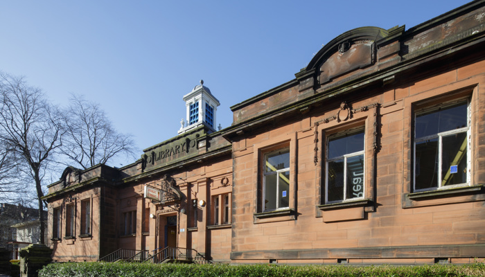 An exterior close-up image of Langside Library which is a red sandstone building built in 1915. There is a 'Library' sign carved into the building above the doorway and also a projecting 'Public Library' sign. There are 3 large windows at each side of the entrance. There are hedges surrounding the front of the building. This photograph has been taken on a sunny day in winter - you can see a bare tree in the background.