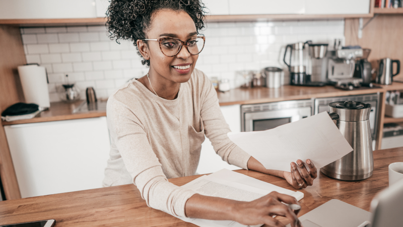 A person is working on a laptop from their kitchen. They have papers in their hand and smiling towards the screen.