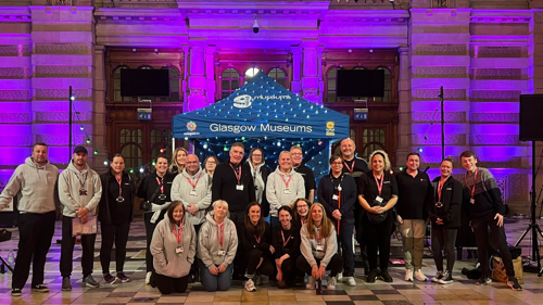 A large group of colleagues gathered for a photo in the grand marble entrance hall of Kelvingrove museum. They are in from of a blue gazebo that is branded Glasgow Museums