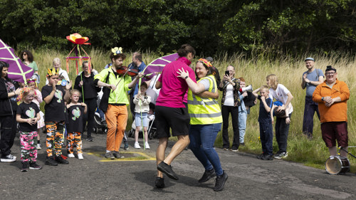 A multi-generational crowd look on as two people dance together, accompanied by a violinist 