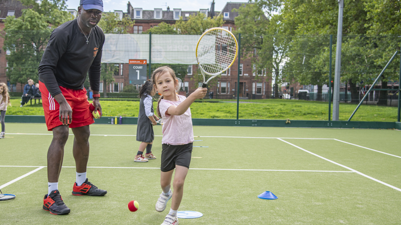 A child swings a tennis racket while a coach stands by watching over. They are at an outdoor grass tennis court.