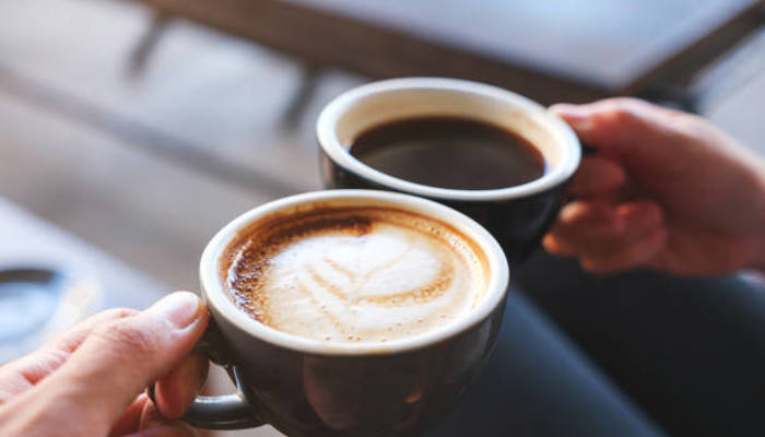 Image of two hands holding coffee cups. One is holding a black coffee and one is holding a coffee with foamy milk. The background is blurred but you can make out a table, likely a coffee shop.