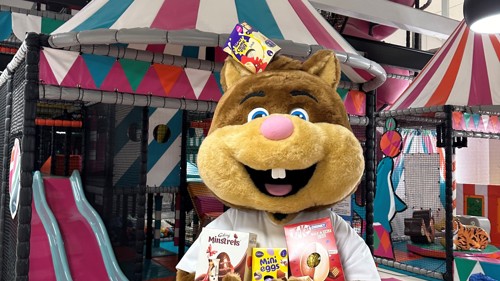A person in a smiling squirrel mascot costume is holding a bunch of chocolate Easter eggs while standing in front of a colourful indoor soft play area