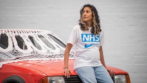 Artist Jasleen Kaur sitting on the bonnet of an old red Ford Escort with a very large doily draped over the roof. They are wearing jeans and a white t-shirt with NHS in blue letters on it
