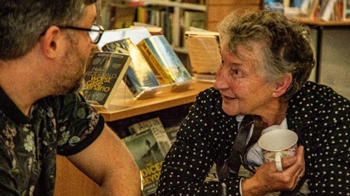 A adult and an elderly person sit in a public library chatting.
