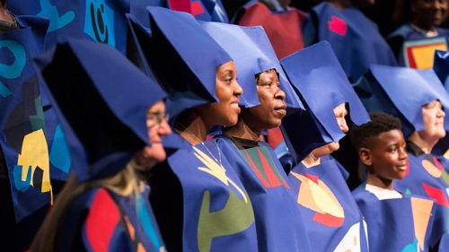 A choir sings, they are wearing creative costumes and hats which are purple.