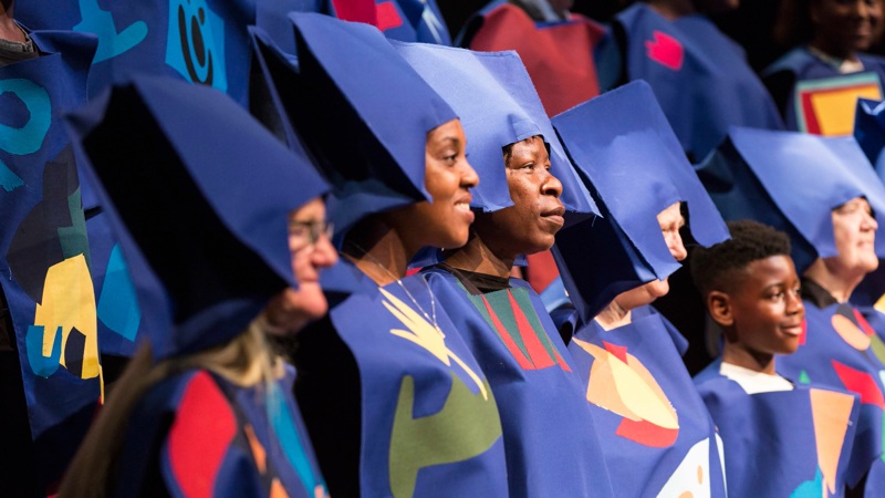 A choir sings, they are wearing creative costumes and hats which are purple.