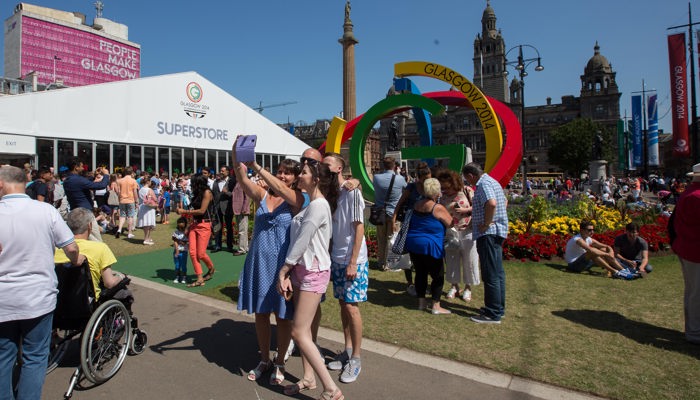 People taking photoes in George Square during the Glasgow 2014 Commonwealth Games. Crowds are gathered outside the superstore and around the Big G sculpture, the Met Tower with People Make Glasgow on it is overlooking the square.