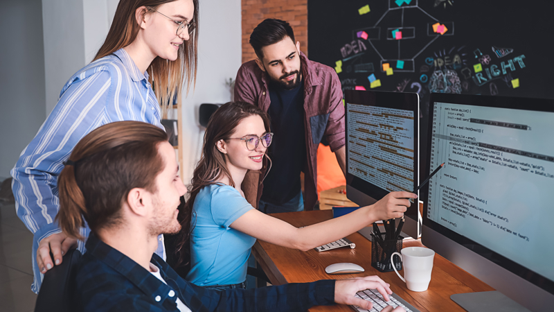 A group of four students are gathered around one computer screen working on a project together. One is pointing at the screen.