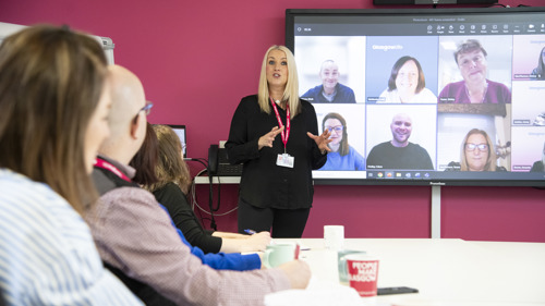 A group of 4 people taking part in a meeting in a meeting room with several people joining online on a large screen. One person is standing at the front of the room talking to the group