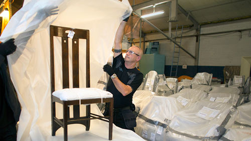 Two people unwrapping plastic from a restored Charles Rennie Mackintosh chair. The image is taken in a warehouse with a number of other items wrapped in plastic in the background.