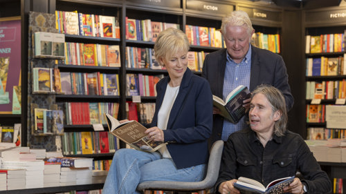 Two men and a woman smile at each other in a library setting, with books in the background and foreground