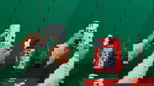 Hands hold a white mug decorated with colorful holiday string lights against a green background, while another decorated mug sits in a red gift bag beside small decorative Christmas trees and crafting supplies like scissors and paper.