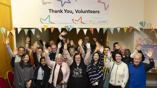 Photo of a large group of people indoors standing under a sign "thank you volunteers" looking at the camera