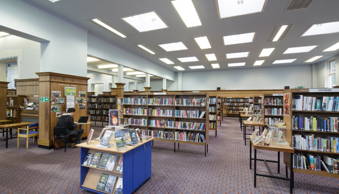 View of the main reading area. There are bookshelves around the perimeter of the room as well and free-standing bookshelves. There are also several tables with book displays. helves in the middle. There is an adult sitting at a small table using their laptop. The room has wood pannelling around a third of the height of the wall and the top of the walls are painted white.