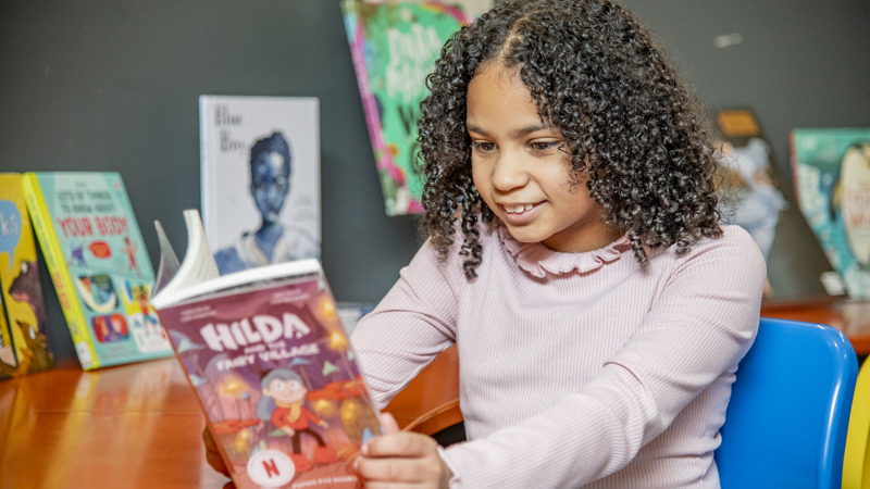 A child smiles reading a book, which has the title ‘Hilda’. Books are displayed on the wall in the background in what is a colourful library scene.
