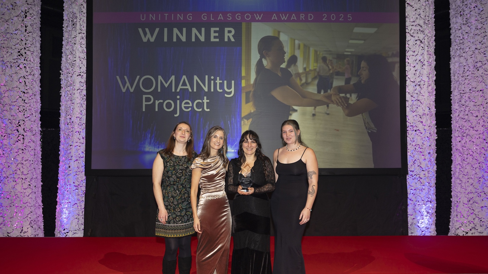 Four people in formal wear stand in a row in front of a big screen which displays the words 'Uniting Glasgow Award 2025 winner' and 'The WOMANity Project'