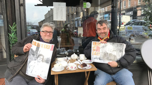 Two people sit in front of a cafe in the East End of Glasgow. The both hole portraits of people with the words Taels Fea the East written on them
