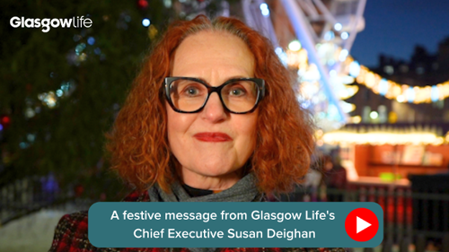 A still from a video showing a person wearing large glasses, a winter coat and scarf standing in front of a lit Christmas tree in a city square 