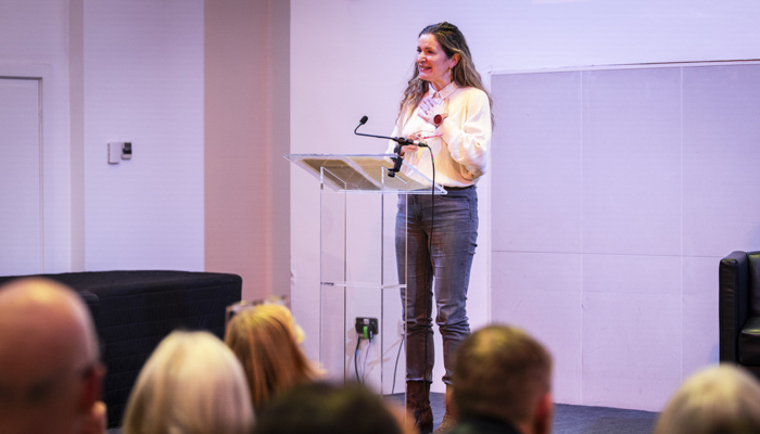 A woman with long hair is standing at a clear podium, speaking into a microphone during an event.