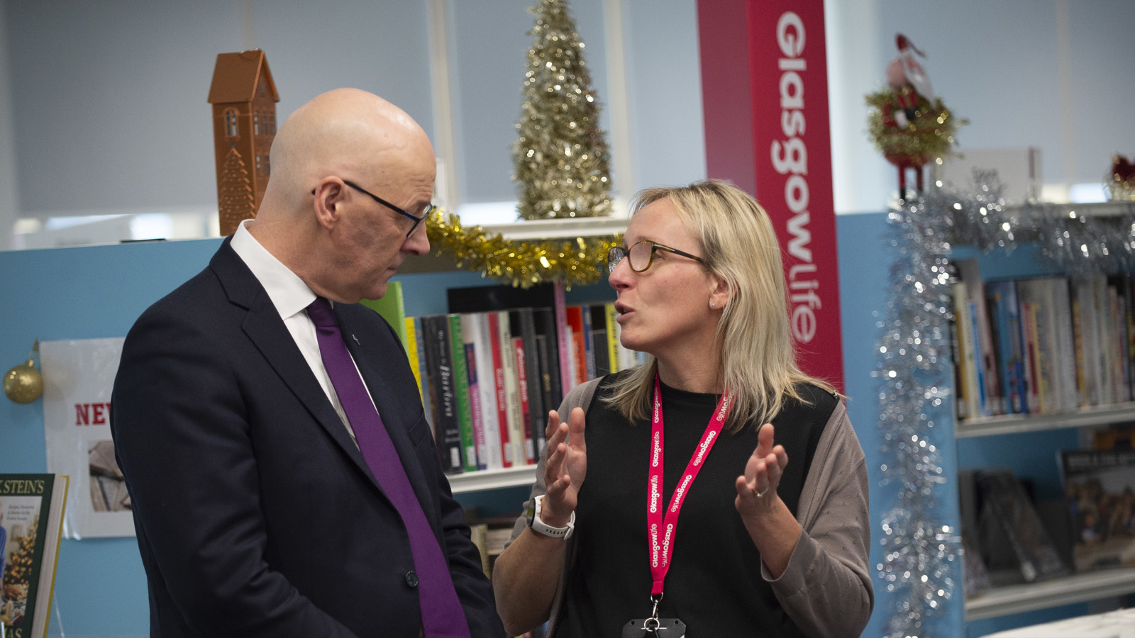 In a decorated library space with bookshelves and tinsel, a bald man in a suit listens while a woman wearing glasses and a staff lanyard gestures with both hands as she speaks. A small Christmas tree and various books are visible behind them.