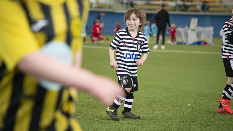 A child, wearing a black and white football strip, smiles widely while on a grassy football pitch. Other players, who are blurry, can be seen dotted around the pitch.