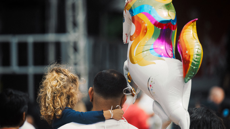 A large unicorn balloon floats above the heads of an adult and child, with the string of the balloon attached to the child's wrist.