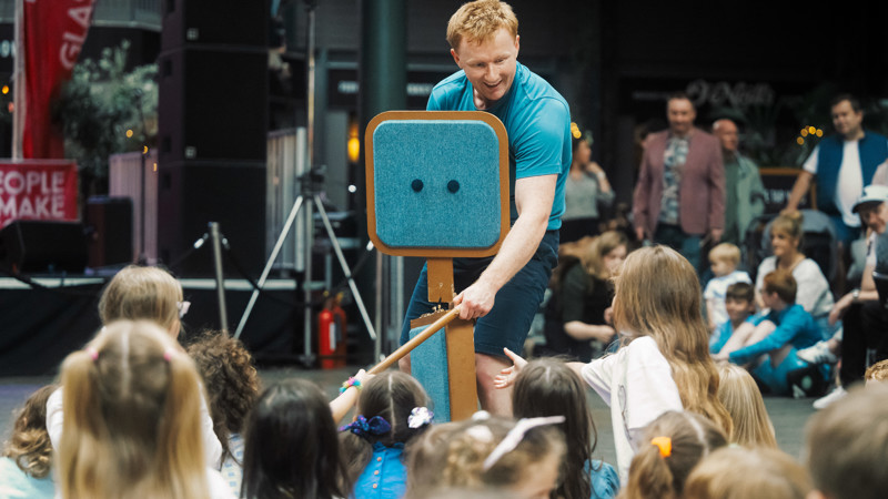 A performer on a small stage, wearing a bold blue tee-shirt, performs in front of a crowd of children. 