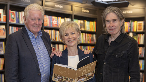 Two men and a woman stand in a classically furnished library. The woman is standing in the middle of the three holding a book