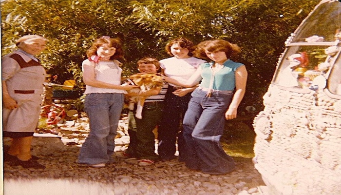 Flora is shown on the left wearing an overall and has grey hair and glasses. To the right are three adults and a younger child holding a dog. Everyone is smiling.