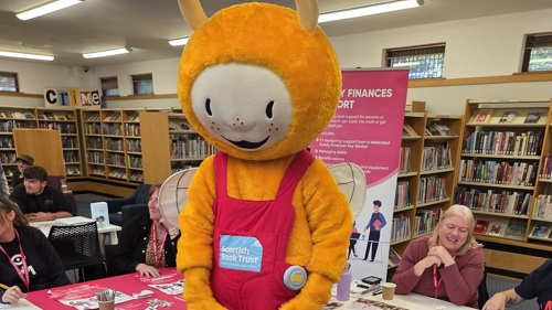 A large yellow mascot character is entertaining a group of young people in a library. There are information stalls and shelves of books behind