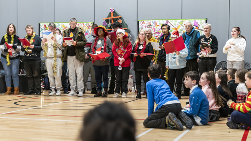 A group of people of various ages sing as choir in a school hall.