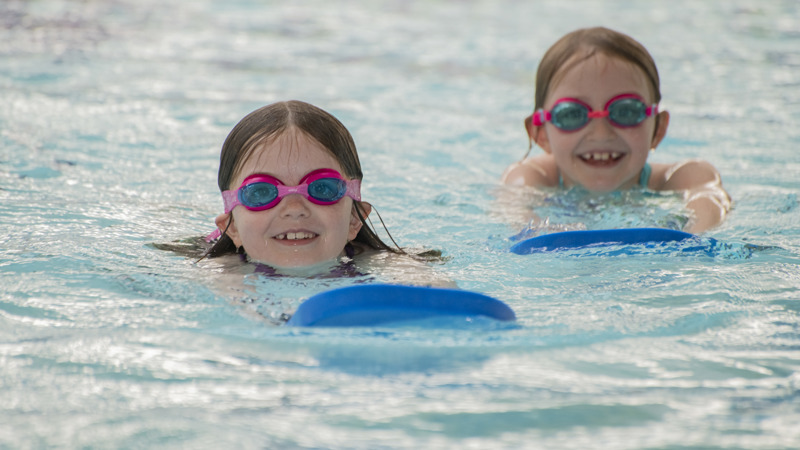 Two children’s heads bob above the water in a swimming pool. Both are smiling widely, holding blue swim floats and wearing pink goggles. 