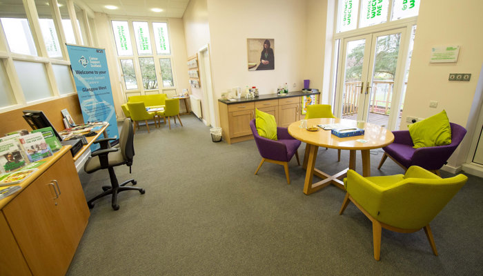 Health and Wellbeing area with 2 tables surrounded by soft purple and green chairs. There is also a desk with a PC at the side of the room. The room has large, tall windows and there are patio doors near one of the tables