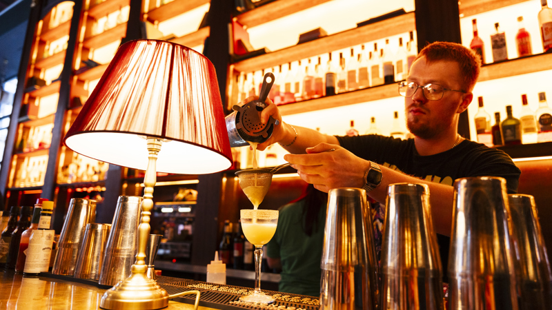 Bartender pouring a cocktail at a bar with warm lighting, shelves of bottles in the background.