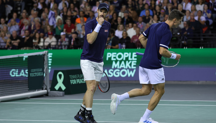 Andy Murray on court clenching his fist during the Davis Cup at the Emirates Arena in 2022, behind him and his partner is a People Make Glasgow advertising hoarding.