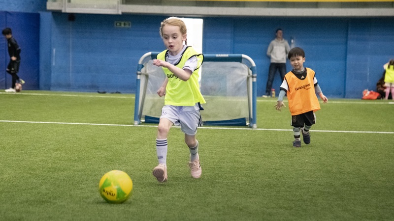 A child in a yellow bib is running towards a football and about to strike it in an indoor pitch. A younger child in an orange bib is in the background on the pitch.