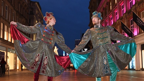 Two performers in glittery regal costumes and wearing roller skates posing for a photo on a pedestrianised shopping street in the evening