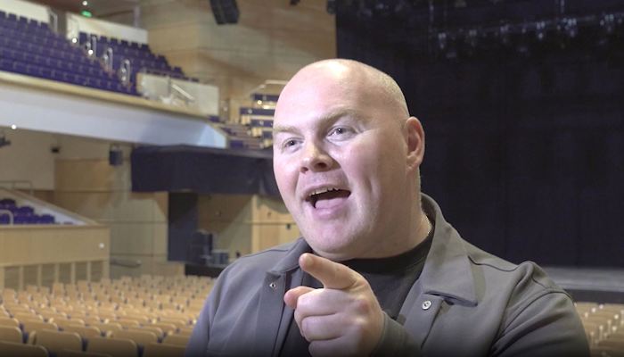 A bald man in a dark jacket smiles and points while seated in an empty theatre.