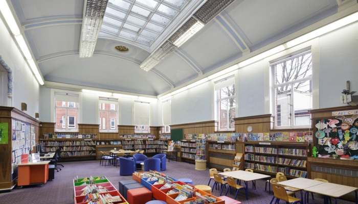 A large spacios childrens area which is long and rectangular. There are wooden bookshelves filled with colourful children's books along the perimeter of the room. There is a high curved ceiling with a long grid-colonial style skylight. There is wooden panelling around a third of the walls. The rest of the walls and ceiling are painted white. There is also colourful children's artwork on the walls, tables and chairs and colourful book storage boxes in the middle of the room.