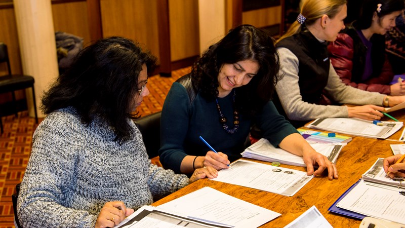 4 women from different ethnic backgrounds sitting at a desk with books, pens and paper in front of them
