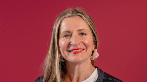 A woman with light brown hair smiles in front of a rubine background